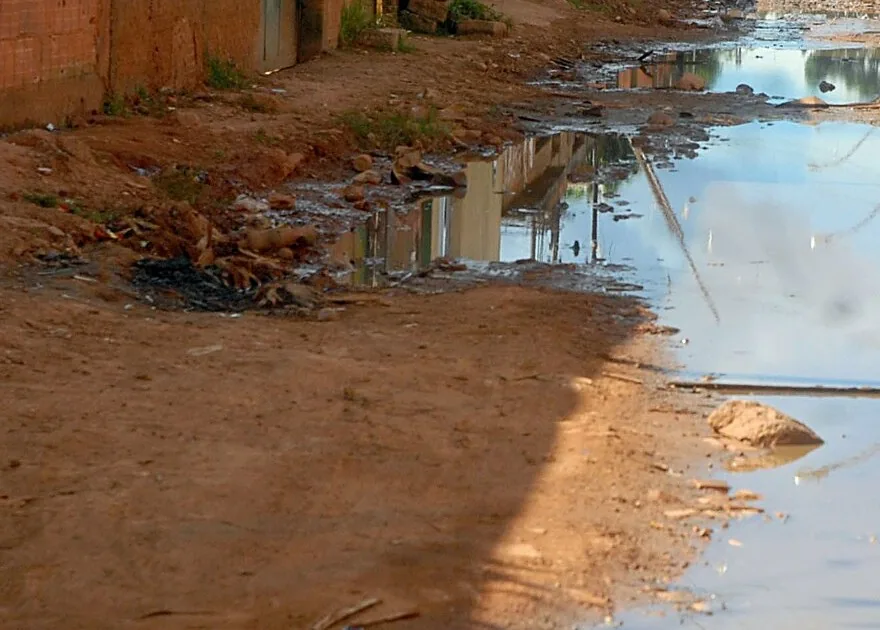 Rua de terra com esgoto correndo a céu aberto entre casas de tijolo